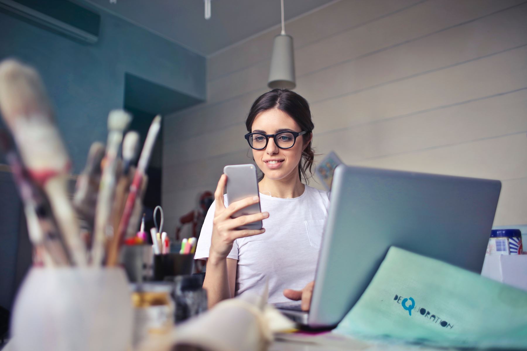 woman holding and looking at a cell phone while working on a laptop